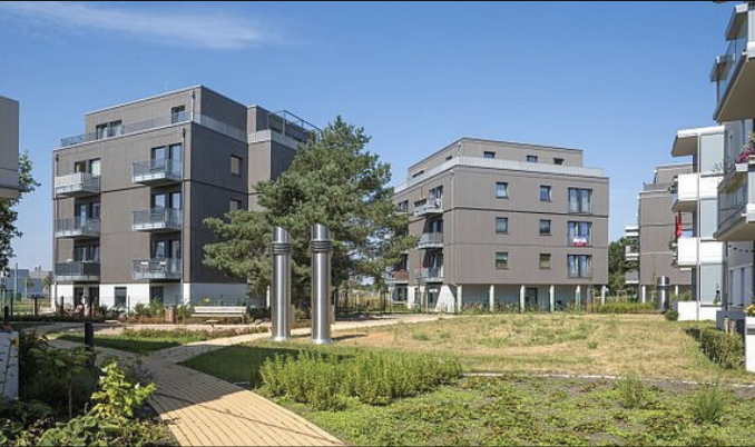 Moderne Wohngebäude mit Balkonen, begrüntem Innenhof und Holzweg unter blauem Himmel